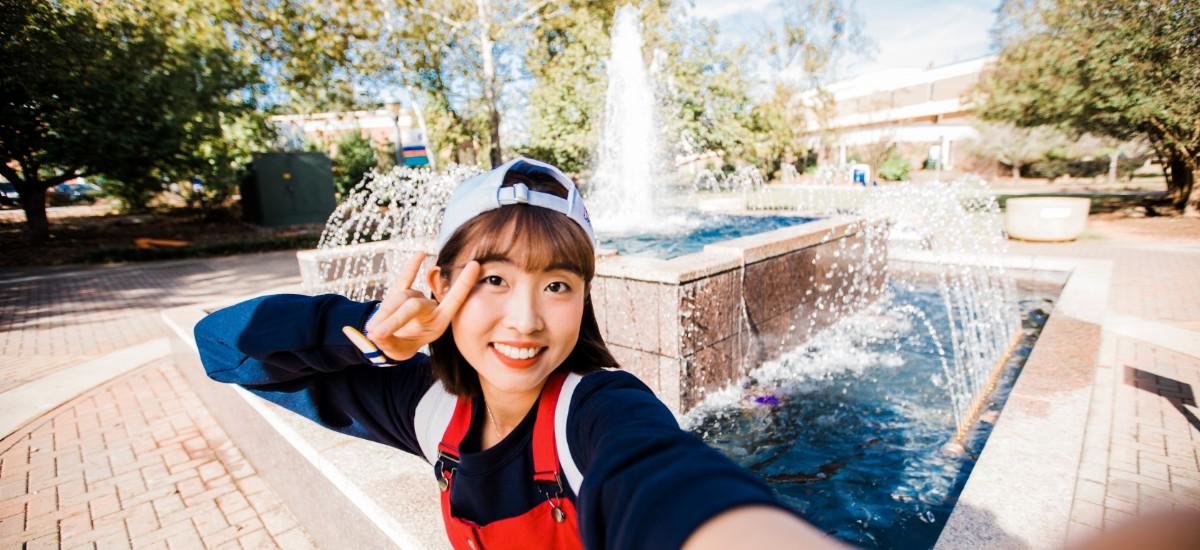 Female student showing the wolf sign in front of the campus fountain.